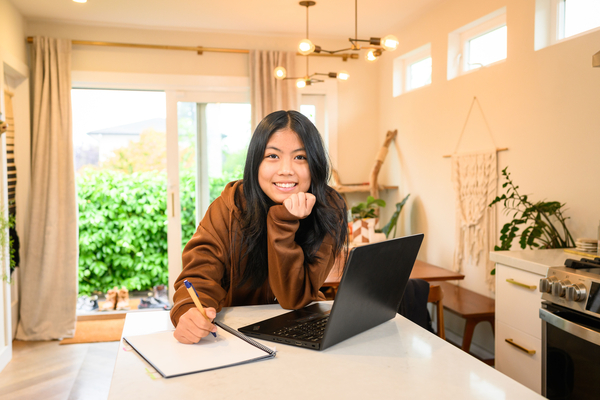 Asian young adult in from of a lap top writting in a notebook